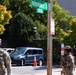 Two armed military personnel in camouflage uniforms are stationed at the intersection of Independence Avenue SW at the 900 block, likely in Washington D.C. They appear to be providing security at what looks like a controlled access point, with a traffic cone blocking vehicle entry. A civilian with a badge/lanyard is visible in the background near a building entrance.