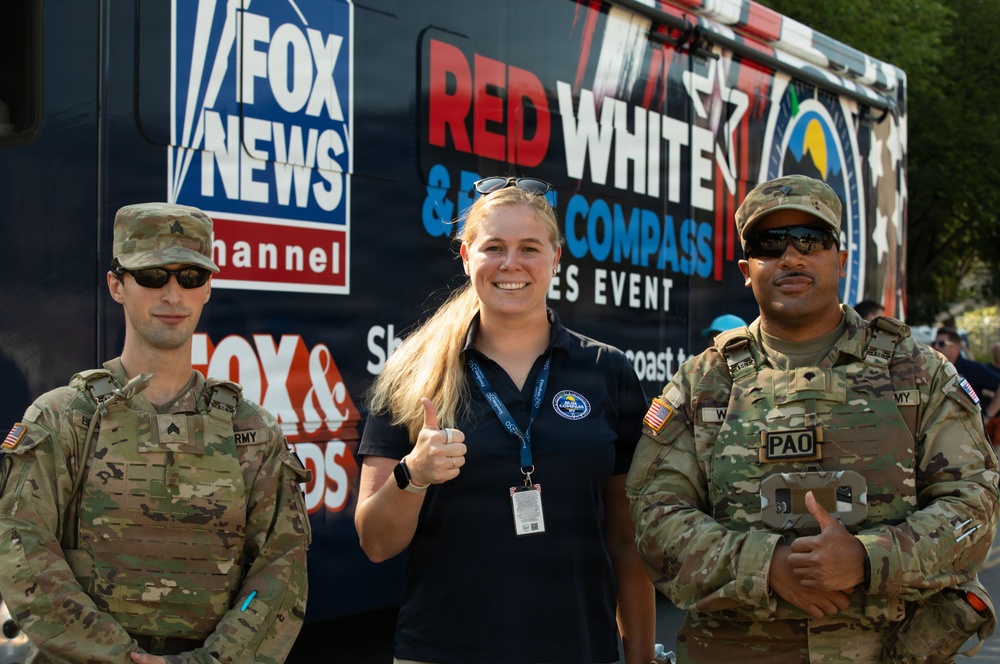 Three individuals pose in front of a Fox News Channel vehicle at what appears to be a 'Red White &amp; Blue Compass Series Event.' Two U.S. Army soldiers in MultiCam camouflage uniforms flank a civilian woman wearing a dark polo shirt with a lanyard and credential badge. The soldier on the right has a 'PAO' (Public Affairs Officer) patch visible on his uniform.