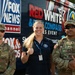 Three individuals pose in front of a Fox News Channel vehicle at what appears to be a 'Red White &amp; Blue Compass Series Event.' Two U.S. Army soldiers in MultiCam camouflage uniforms flank a civilian woman wearing a dark polo shirt with a lanyard and credential badge. The soldier on the right has a 'PAO' (Public Affairs Officer) patch visible on his uniform.