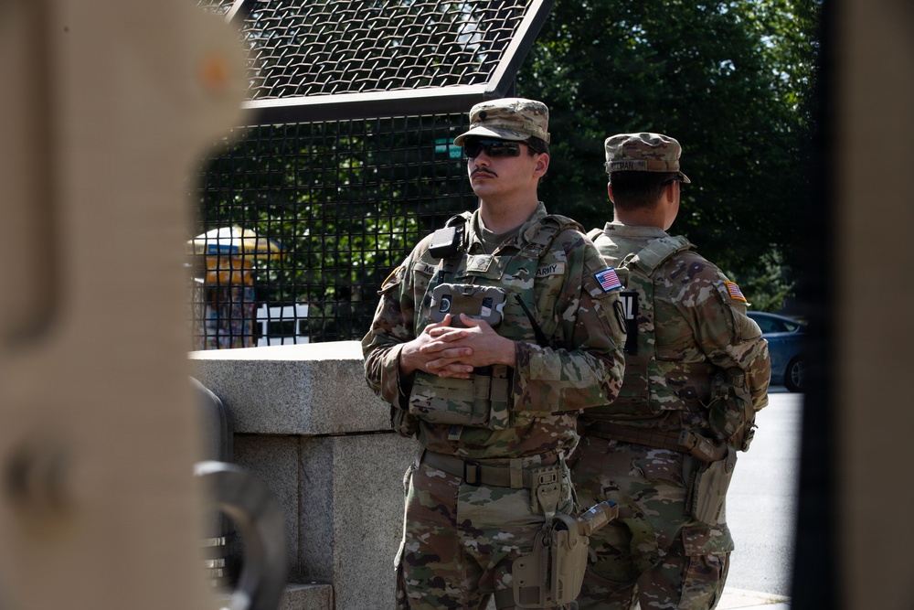 Two U.S. Army soldiers in full combat uniforms and tactical gear are standing at what appears to be a security post or checkpoint. The soldier facing the camera wears sunglasses, a patrol cap, body armor, and has an 'ARMY' tape and U.S. flag patch visible on his uniform. They appear to be on guard duty near a barrier or fencing structure.
