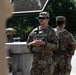 Two U.S. Army soldiers in full combat uniforms and tactical gear are standing at what appears to be a security post or checkpoint. The soldier facing the camera wears sunglasses, a patrol cap, body armor, and has an 'ARMY' tape and U.S. flag patch visible on his uniform. They appear to be on guard duty near a barrier or fencing structure.