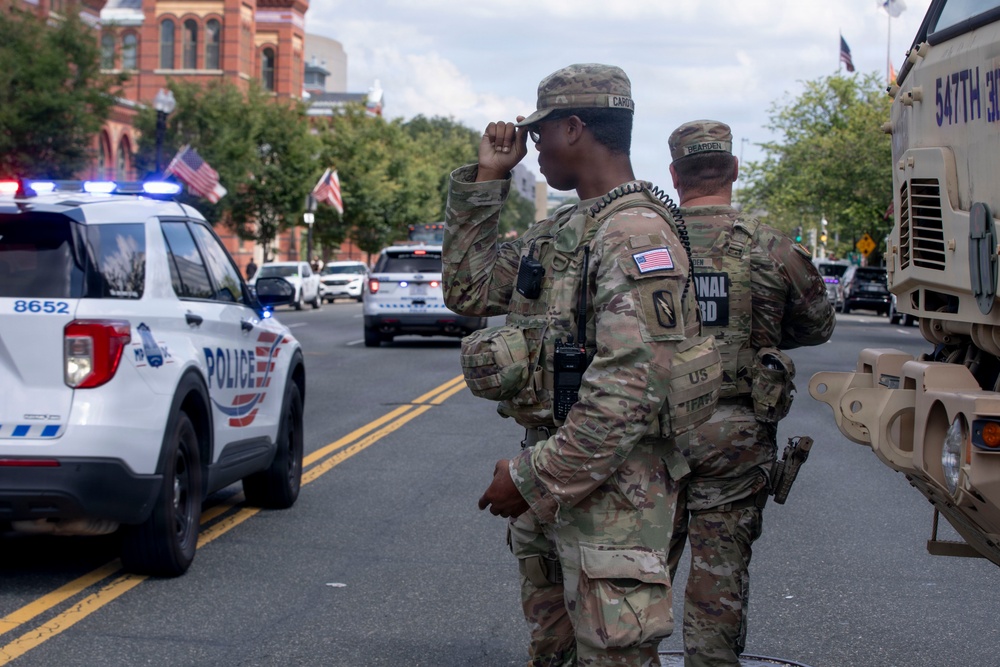 Two U.S. National Guard soldiers in MultiCam camouflage uniforms are standing on a street alongside a Washington D.C. Metropolitan Police vehicle (unit 8652). A military vehicle marked '547th' is visible to their right. The scene suggests a security or crowd control operation in an urban Washington D.C. setting.