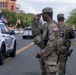 Two U.S. National Guard soldiers in MultiCam camouflage uniforms are standing on a street alongside a Washington D.C. Metropolitan Police vehicle (unit 8652). A military vehicle marked '547th' is visible to their right. The scene suggests a security or crowd control operation in an urban Washington D.C. setting.