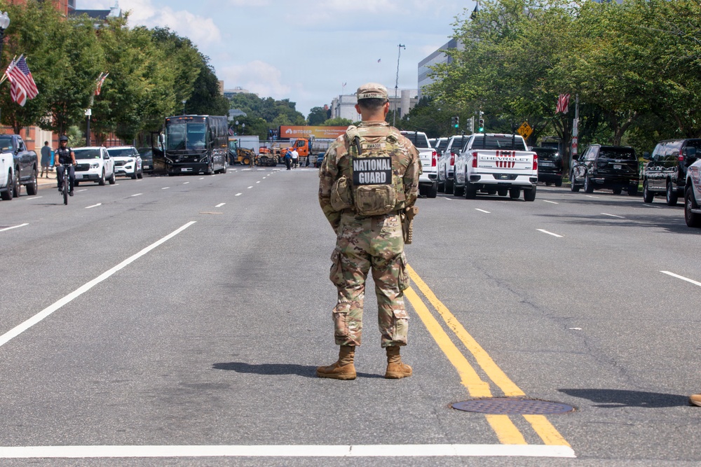 A National Guard soldier stands in the center of a divided urban street, facing away from the camera, appearing to monitor or control traffic and crowd movement. The scene suggests a security or law enforcement operation, with multiple police vehicles, civilian vehicles, and personnel visible along both sides of the street. American flags are visible on light poles, and the setting appears to be a major city boulevard, consistent with Washington D.C.