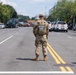 A National Guard soldier stands in the center of a divided urban street, facing away from the camera, appearing to monitor or control traffic and crowd movement. The scene suggests a security or law enforcement operation, with multiple police vehicles, civilian vehicles, and personnel visible along both sides of the street. American flags are visible on light poles, and the setting appears to be a major city boulevard, consistent with Washington D.C.