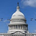 The image shows the United States Capitol Building in Washington, D.C., with its iconic white dome and the Statue of Freedom at its apex. Multiple military helicopters are flying in formation on both sides of the dome against a partly cloudy blue sky. This appears to be a military flyover, likely associated with a ceremonial or national event.