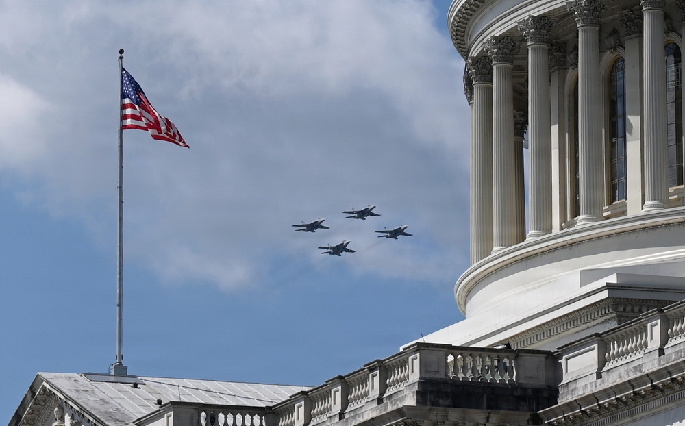 Military jet aircraft fly in formation over a neoclassical government building, identifiable as the United States Capitol by its distinctive dome and architectural style. An American flag flies on a flagpole in the foreground against a partly cloudy blue sky. The scene appears to depict a ceremonial flyover, likely associated with a national event or commemoration.
