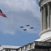 Military jet aircraft fly in formation over a neoclassical government building, identifiable as the United States Capitol by its distinctive dome and architectural style. An American flag flies on a flagpole in the foreground against a partly cloudy blue sky. The scene appears to depict a ceremonial flyover, likely associated with a national event or commemoration.
