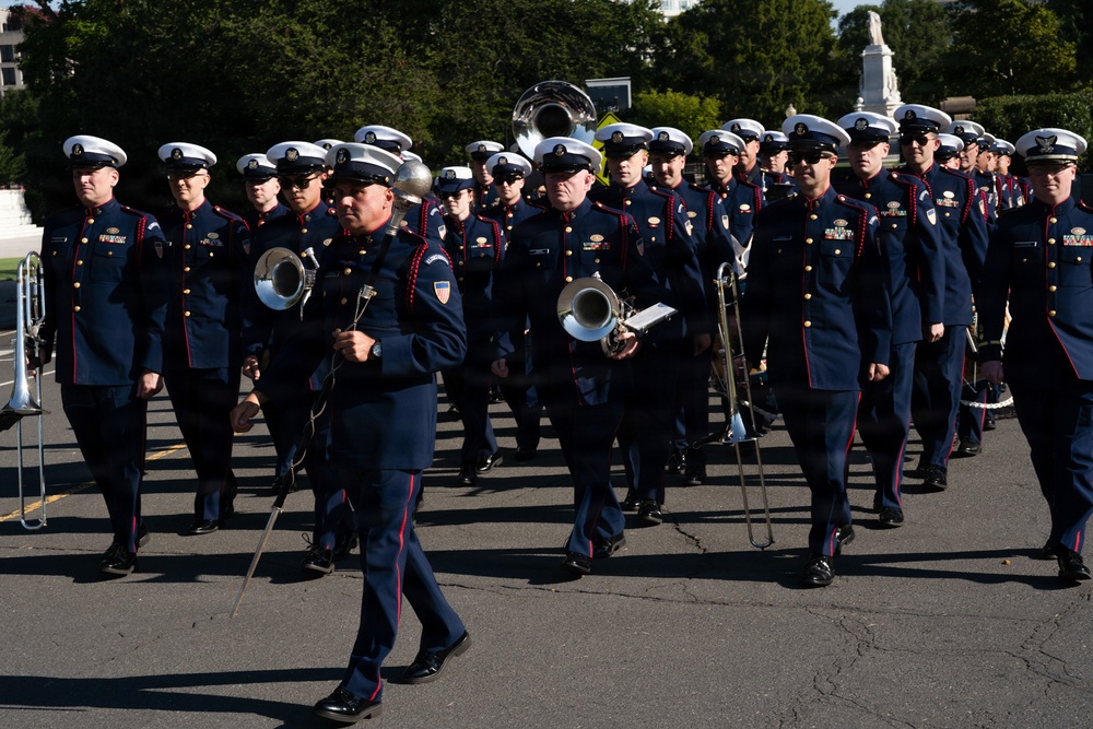 A military marching band is parading in formation along a paved road, wearing dark navy dress uniforms with red trim and white peaked caps. The musicians are carrying brass instruments including trombones, sousaphones, and bugles. The setting appears to be a formal ceremonial event, likely in Washington D.C., based on the visible monument and architecture in the background.