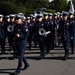 A military marching band is parading in formation along a paved road, wearing dark navy dress uniforms with red trim and white peaked caps. The musicians are carrying brass instruments including trombones, sousaphones, and bugles. The setting appears to be a formal ceremonial event, likely in Washington D.C., based on the visible monument and architecture in the background.