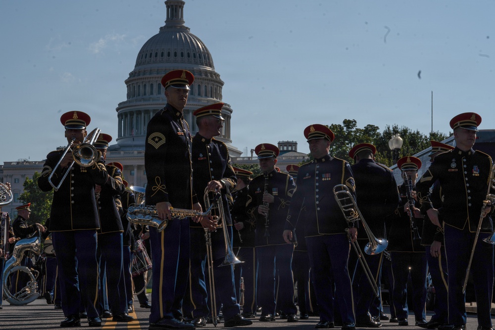 A U.S. Army military band is performing or assembling in formation on a street directly in front of the United States Capitol building in Washington, D.C. The musicians are dressed in formal dark blue and black dress uniforms with distinctive red-topped ceremonial caps and gold trim. The scene appears to be part of an official military ceremony or parade event.
