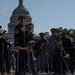 A U.S. Army military band is performing or assembling in formation on a street directly in front of the United States Capitol building in Washington, D.C. The musicians are dressed in formal dark blue and black dress uniforms with distinctive red-topped ceremonial caps and gold trim. The scene appears to be part of an official military ceremony or parade event.