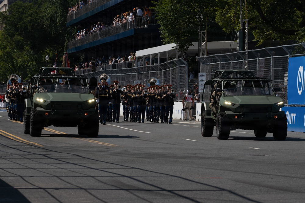 A military parade is taking place on a wide urban street, featuring two olive-green military utility vehicles (likely Infantry Squad Vehicles or similar light tactical vehicles) flanking a marching military band in dress uniforms. Spectators are watching from multi-level grandstands in the background. The setting appears to be a formal ceremonial parade route with security fencing and sponsor signage visible.