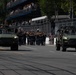 A military parade is taking place on a wide urban street, featuring two olive-green military utility vehicles (likely Infantry Squad Vehicles or similar light tactical vehicles) flanking a marching military band in dress uniforms. Spectators are watching from multi-level grandstands in the background. The setting appears to be a formal ceremonial parade route with security fencing and sponsor signage visible.