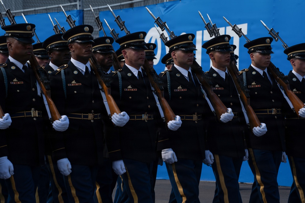 A formation of U.S. Army soldiers in dress uniform are marching in a parade or ceremonial event. They are carrying rifles with fixed bayonets and wearing formal black service uniforms with white gloves. The soldiers are moving in tight, synchronized formation past a blue banner backdrop.