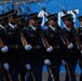 A formation of U.S. Army soldiers in dress uniform are marching in a parade or ceremonial event. They are carrying rifles with fixed bayonets and wearing formal black service uniforms with white gloves. The soldiers are moving in tight, synchronized formation past a blue banner backdrop.