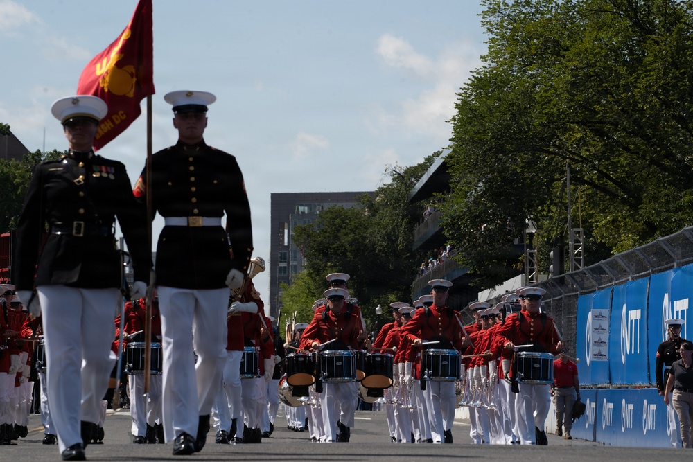 A U.S. Marine Corps marching band and color guard are performing in an outdoor parade along a city street. The band members are dressed in red tunics with white trousers, while the color guard wears the traditional dress blue uniform. A red Marine Corps flag is prominently carried at the front of the procession.