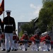 A U.S. Marine Corps marching band and color guard are performing in an outdoor parade along a city street. The band members are dressed in red tunics with white trousers, while the color guard wears the traditional dress blue uniform. A red Marine Corps flag is prominently carried at the front of the procession.