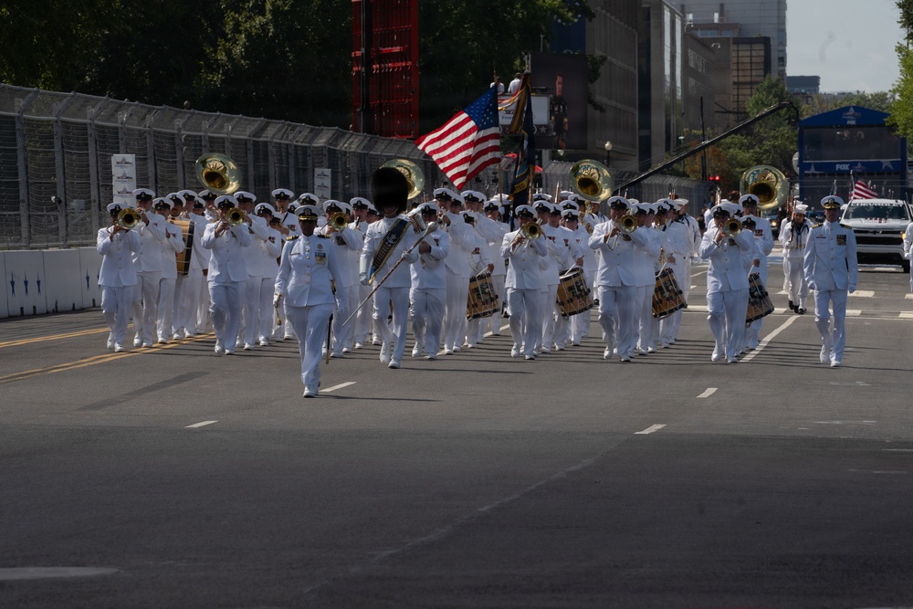 A military marching band dressed in white dress uniforms is parading down a closed urban street. The band includes brass instruments, drums, and a drum major leading the formation, with an American flag and what appears to be a unit colors flag visible in the background. The parade takes place in an urban setting with buildings, trees, and temporary barriers lining the route.
