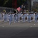 A military marching band dressed in white dress uniforms is parading down a closed urban street. The band includes brass instruments, drums, and a drum major leading the formation, with an American flag and what appears to be a unit colors flag visible in the background. The parade takes place in an urban setting with buildings, trees, and temporary barriers lining the route.