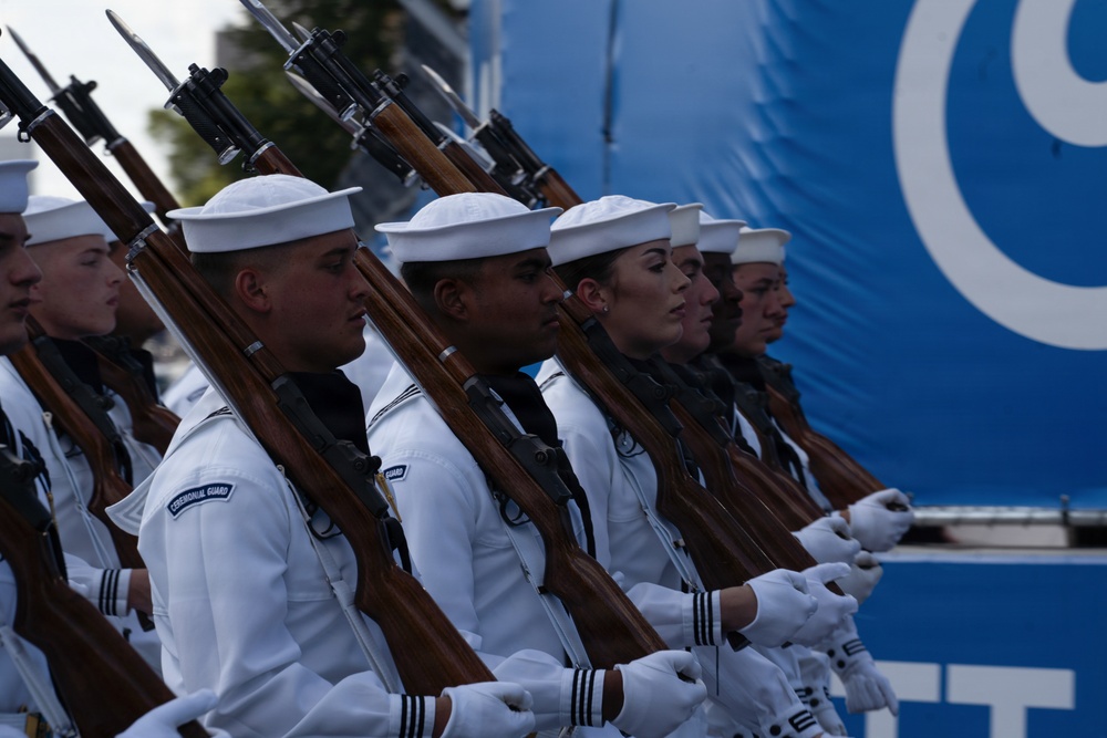A formation of U.S. Navy Ceremonial Guard members stands at attention during a formal parade or ceremony. Personnel are dressed in white dress uniforms and white gloves, carrying rifles with fixed bayonets at a uniform angle. The group includes both male and female sailors marching in precise alignment.