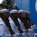 A formation of U.S. Navy Ceremonial Guard members stands at attention during a formal parade or ceremony. Personnel are dressed in white dress uniforms and white gloves, carrying rifles with fixed bayonets at a uniform angle. The group includes both male and female sailors marching in precise alignment.