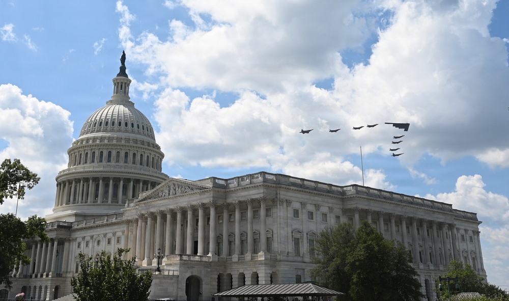 The image shows the United States Capitol Building in Washington, D.C., photographed from a low angle on a partly cloudy day. A formation of military aircraft is visible flying over the building, suggesting a flyover event such as a national holiday or ceremonial occasion. The neoclassical architecture of the Capitol, including its iconic dome topped with the Statue of Freedom, is clearly visible.