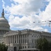 The image shows the United States Capitol Building in Washington, D.C., photographed from a low angle on a partly cloudy day. A formation of military aircraft is visible flying over the building, suggesting a flyover event such as a national holiday or ceremonial occasion. The neoclassical architecture of the Capitol, including its iconic dome topped with the Statue of Freedom, is clearly visible.