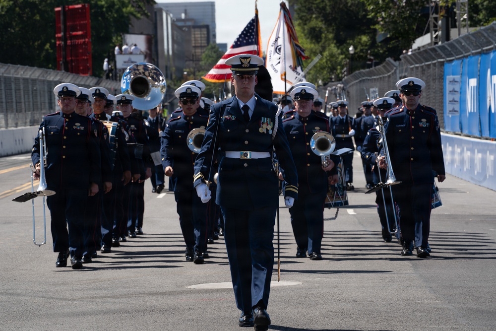 A military marching band parades down a closed street or racing circuit, led by a drum major in dark navy dress uniform carrying a baton. Band members in matching navy uniforms with white caps carry brass instruments including tubas, trombones, and mellophone-style horns. An American flag and additional ceremonial flags are visible being carried in the formation behind the drum major.