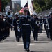 A military marching band parades down a closed street or racing circuit, led by a drum major in dark navy dress uniform carrying a baton. Band members in matching navy uniforms with white caps carry brass instruments including tubas, trombones, and mellophone-style horns. An American flag and additional ceremonial flags are visible being carried in the formation behind the drum major.