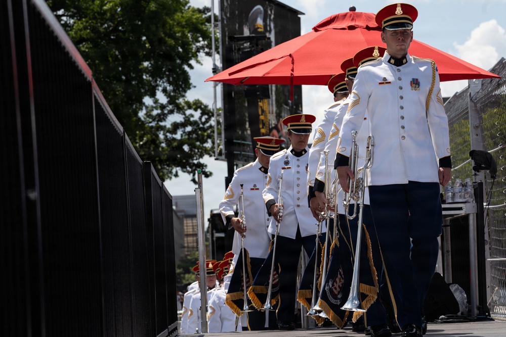 A military band is marching in formation on an outdoor stage or platform, wearing formal white dress uniforms with red caps and gold accents. The musicians are carrying silver herald trumpets with decorative gold-fringed banners. The setting appears to be an outdoor ceremonial or public performance event.