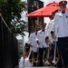 A military band is marching in formation on an outdoor stage or platform, wearing formal white dress uniforms with red caps and gold accents. The musicians are carrying silver herald trumpets with decorative gold-fringed banners. The setting appears to be an outdoor ceremonial or public performance event.