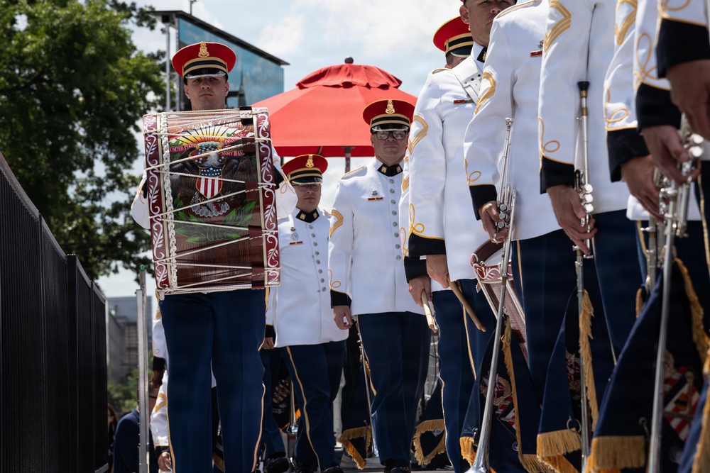 A military marching band, identifiable as the United States Army Band, is shown in formation during what appears to be a formal parade or ceremonial event. The musicians are dressed in distinctive white dress uniforms with gold trim, red-crowned caps, and blue trousers. A prominent bass drum bearing the U.S. Army Band insignia with an eagle emblem is visible in the foreground.