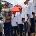 A military marching band, identifiable as the United States Army Band, is shown in formation during what appears to be a formal parade or ceremonial event. The musicians are dressed in distinctive white dress uniforms with gold trim, red-crowned caps, and blue trousers. A prominent bass drum bearing the U.S. Army Band insignia with an eagle emblem is visible in the foreground.