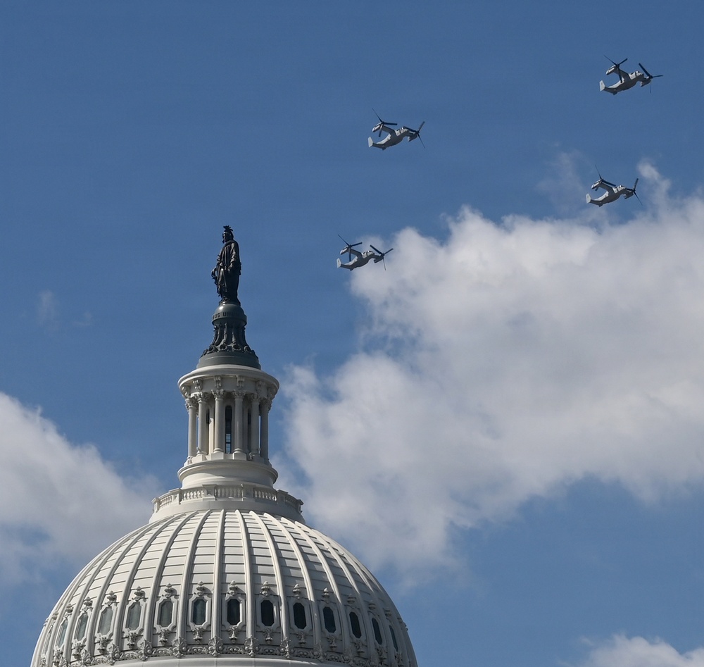 The image shows the dome of the United States Capitol Building with the Statue of Freedom atop it, set against a partly cloudy blue sky. Four military tiltrotor aircraft, identifiable as V-22 Osprey variants, are flying in formation above and to the right of the dome. This appears to be a ceremonial flyover, likely associated with a national event or inauguration.