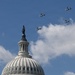 The image shows the dome of the United States Capitol Building with the Statue of Freedom atop it, set against a partly cloudy blue sky. Four military tiltrotor aircraft, identifiable as V-22 Osprey variants, are flying in formation above and to the right of the dome. This appears to be a ceremonial flyover, likely associated with a national event or inauguration.