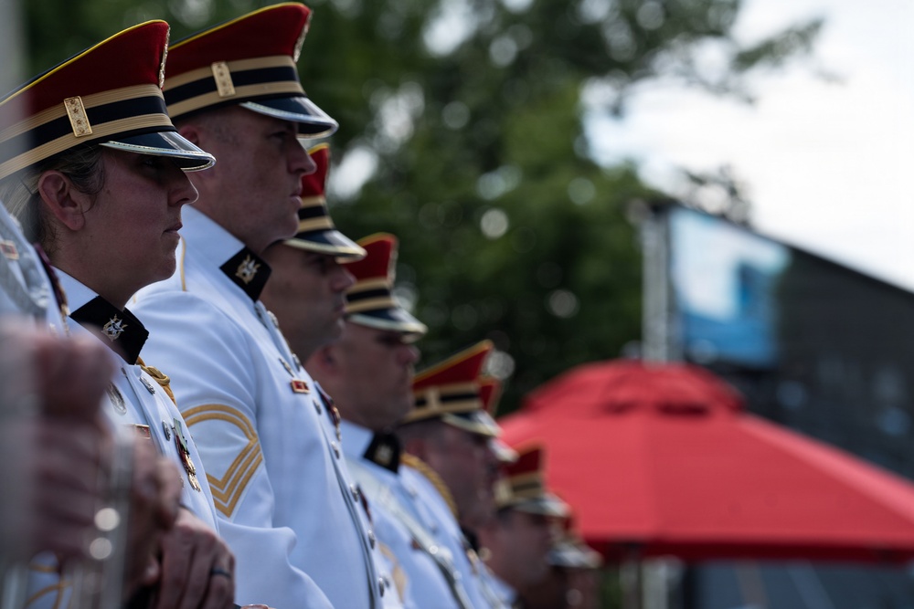 A group of military personnel in formal dress uniforms are standing at attention in a line, facing the same direction. They are wearing distinctive white tunics with gold trim and red-topped ceremonial caps with black visors. The setting appears to be an outdoor ceremonial or parade event.
