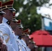 A group of military personnel in formal dress uniforms are standing at attention in a line, facing the same direction. They are wearing distinctive white tunics with gold trim and red-topped ceremonial caps with black visors. The setting appears to be an outdoor ceremonial or parade event.