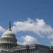 The image shows the United States Capitol Building in Washington, D.C., with its iconic dome and the Statue of Freedom on top. Four military aircraft are flying in formation over the Capitol against a partly cloudy blue sky. The scene appears to capture a ceremonial flyover, likely associated with a national event or commemoration.