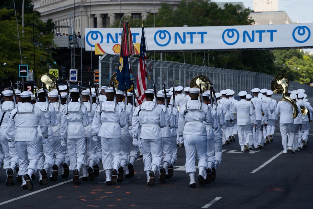 A military parade featuring personnel dressed in white naval uniforms marching in formation down a city street. The procession includes flag bearers carrying the American flag and other military colors, accompanied by a brass marching band with sousaphones. Sponsor banners for NTT and Shell are visible overhead, suggesting this is part of a major public event or race-related ceremony.