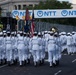A military parade featuring personnel dressed in white naval uniforms marching in formation down a city street. The procession includes flag bearers carrying the American flag and other military colors, accompanied by a brass marching band with sousaphones. Sponsor banners for NTT and Shell are visible overhead, suggesting this is part of a major public event or race-related ceremony.