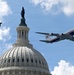 A large military or government Boeing 747-style aircraft with red, white, and blue livery flies over the United States Capitol dome in Washington, D.C. Several smaller military fighter jets are also visible in the sky alongside the large aircraft. The image appears to depict a ceremonial flyover above the Capitol building.