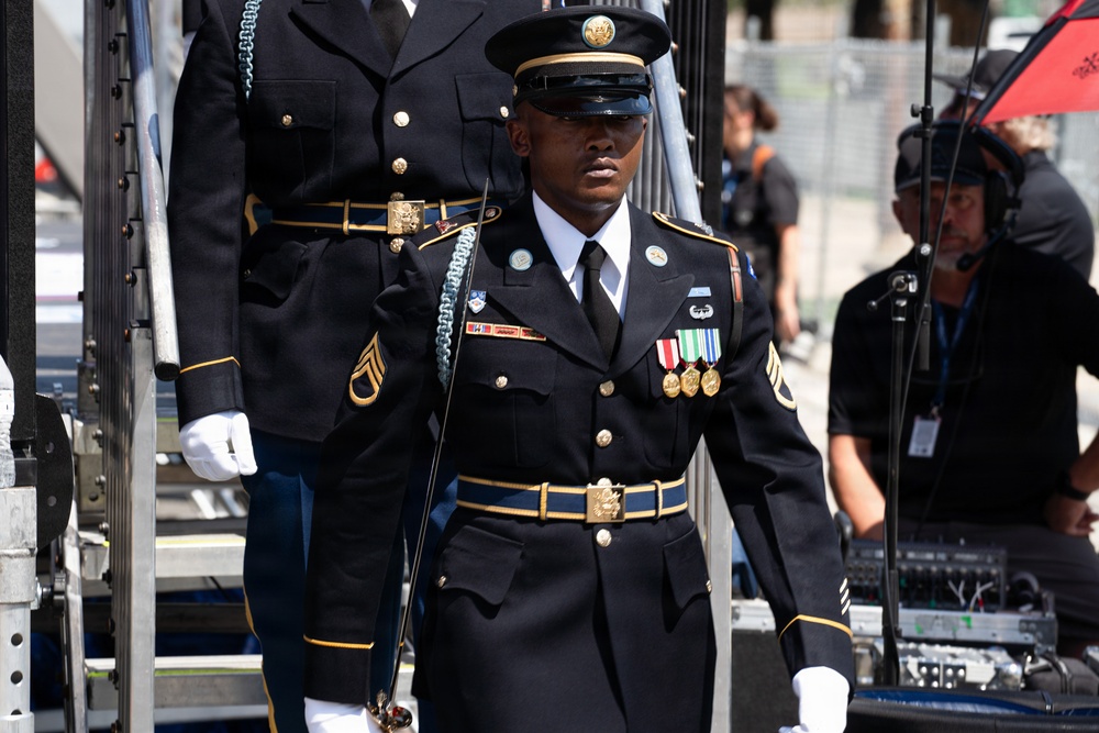 A U.S. Army soldier in formal dress blues uniform stands at attention or marches during what appears to be a ceremonial event or parade. The soldier is wearing multiple service medals, rank insignia consistent with a Staff Sergeant, and is carrying a ceremonial saber. Behind them, another uniformed soldier and civilian event staff with audio equipment are visible.