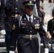 A U.S. Army soldier in formal dress blues uniform stands at attention or marches during what appears to be a ceremonial event or parade. The soldier is wearing multiple service medals, rank insignia consistent with a Staff Sergeant, and is carrying a ceremonial saber. Behind them, another uniformed soldier and civilian event staff with audio equipment are visible.