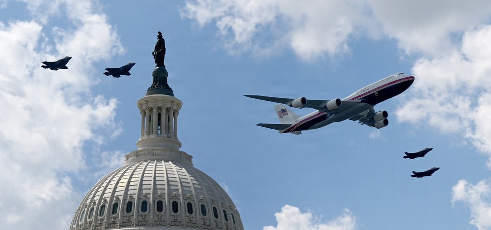 A flyover formation passes above the United States Capitol Building dome in Washington, D.C. The formation includes a large white and red four-engine aircraft bearing U.S. Air Force markings consistent with a VC-25 (Air Force One configuration), accompanied by four smaller fighter jets. The scene appears to be a ceremonial or commemorative aerial flyover.