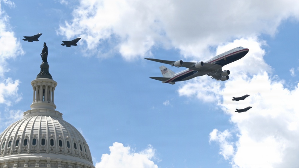 A large Boeing 747 aircraft with blue, white, and red livery bearing a visible American flag marking flies over the United States Capitol building, escorted by four fighter jets. The formation consists of the large aircraft flanked by two fighter jets on each side. The scene appears to be a ceremonial flyover above Washington D.C.
