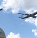 A large Boeing 747 aircraft with blue, white, and red livery bearing a visible American flag marking flies over the United States Capitol building, escorted by four fighter jets. The formation consists of the large aircraft flanked by two fighter jets on each side. The scene appears to be a ceremonial flyover above Washington D.C.
