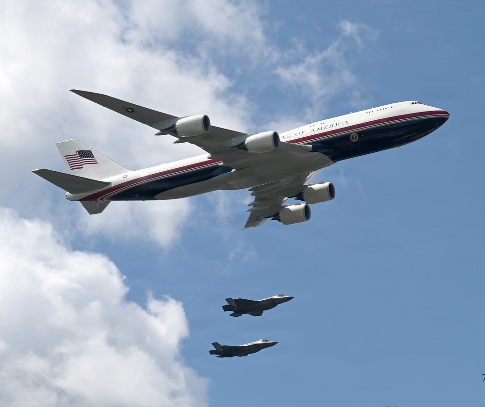 The image shows a large Boeing 747 aircraft bearing the markings 'United States of America' and a Presidential seal, consistent with Air Force One livery, flying in formation with two smaller fighter jets below it. The large aircraft features a white, dark blue, and red color scheme with an American flag on the tail. Two F-35 Lightning II fighter jets appear to be flying in escort formation beneath the larger aircraft against a partly cloudy blue sky.