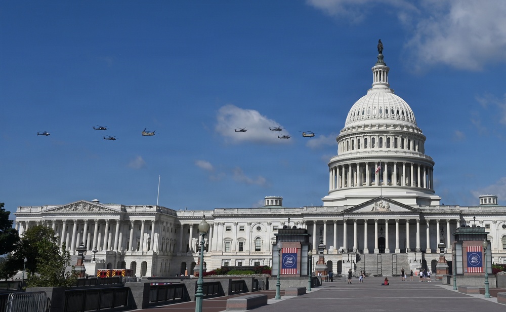 A formation of military helicopters flies over the United States Capitol Building in Washington, D.C. on a clear, sunny day. The Capitol's iconic white dome is prominently visible on the right side of the frame, with decorative banners displayed near the entrance plaza. The scene suggests a ceremonial flyover, likely associated with a national event or celebration.