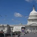 A formation of military helicopters flies over the United States Capitol Building in Washington, D.C. on a clear, sunny day. The Capitol's iconic white dome is prominently visible on the right side of the frame, with decorative banners displayed near the entrance plaza. The scene suggests a ceremonial flyover, likely associated with a national event or celebration.